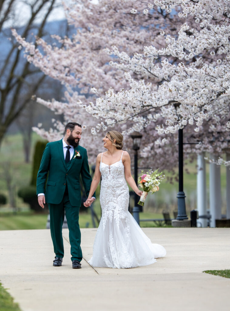 bride in wedding gown and groom in hunter green suit walk under cherry blossom trees
